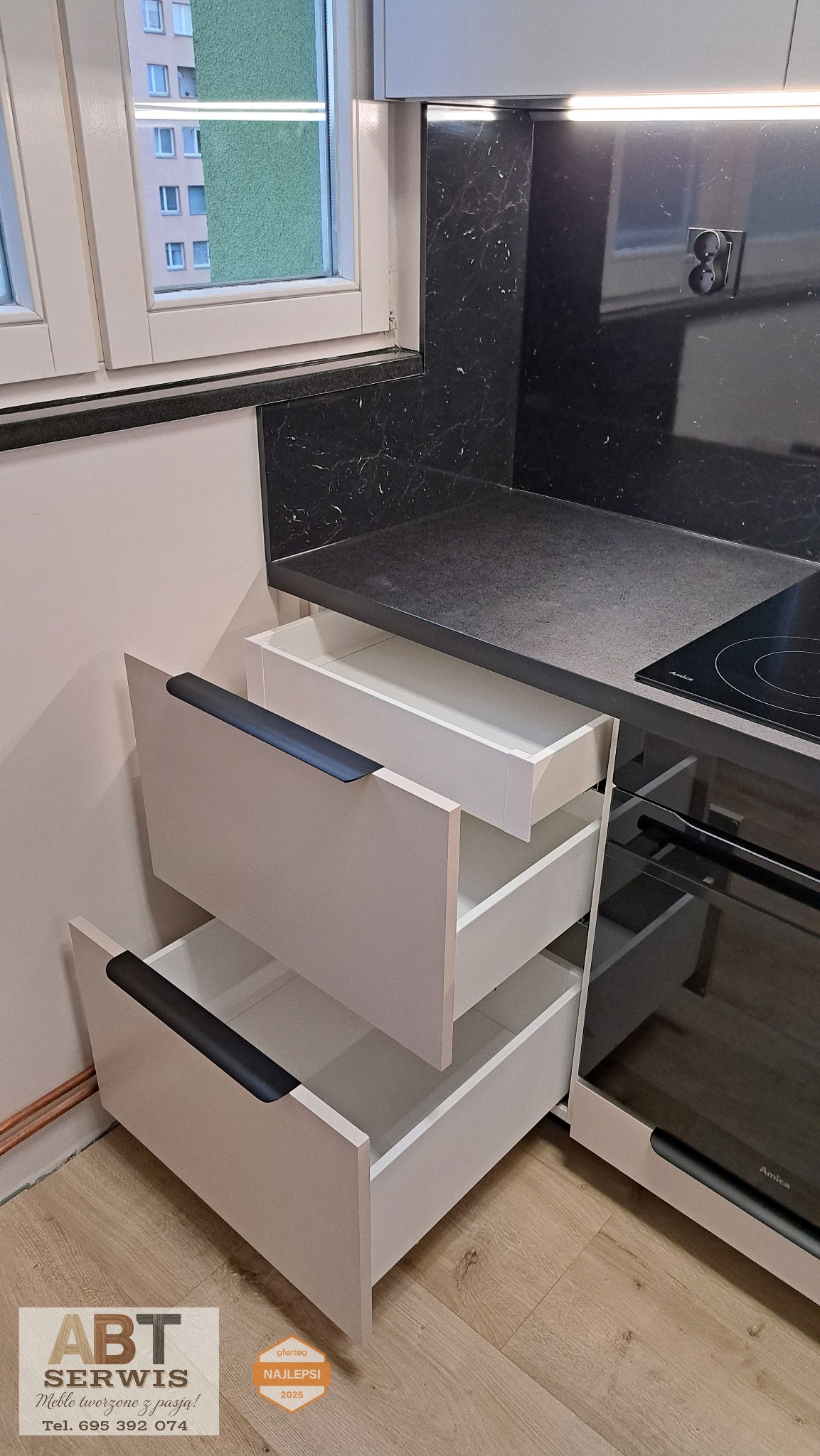 Nowoczesna kuchnia z otwartymi szufladami w odcieniach bieli i szarości, czarny blat z marmurkowym wzorem, płyta indukcyjna Amica, oświetlenie LED pod szafkami.