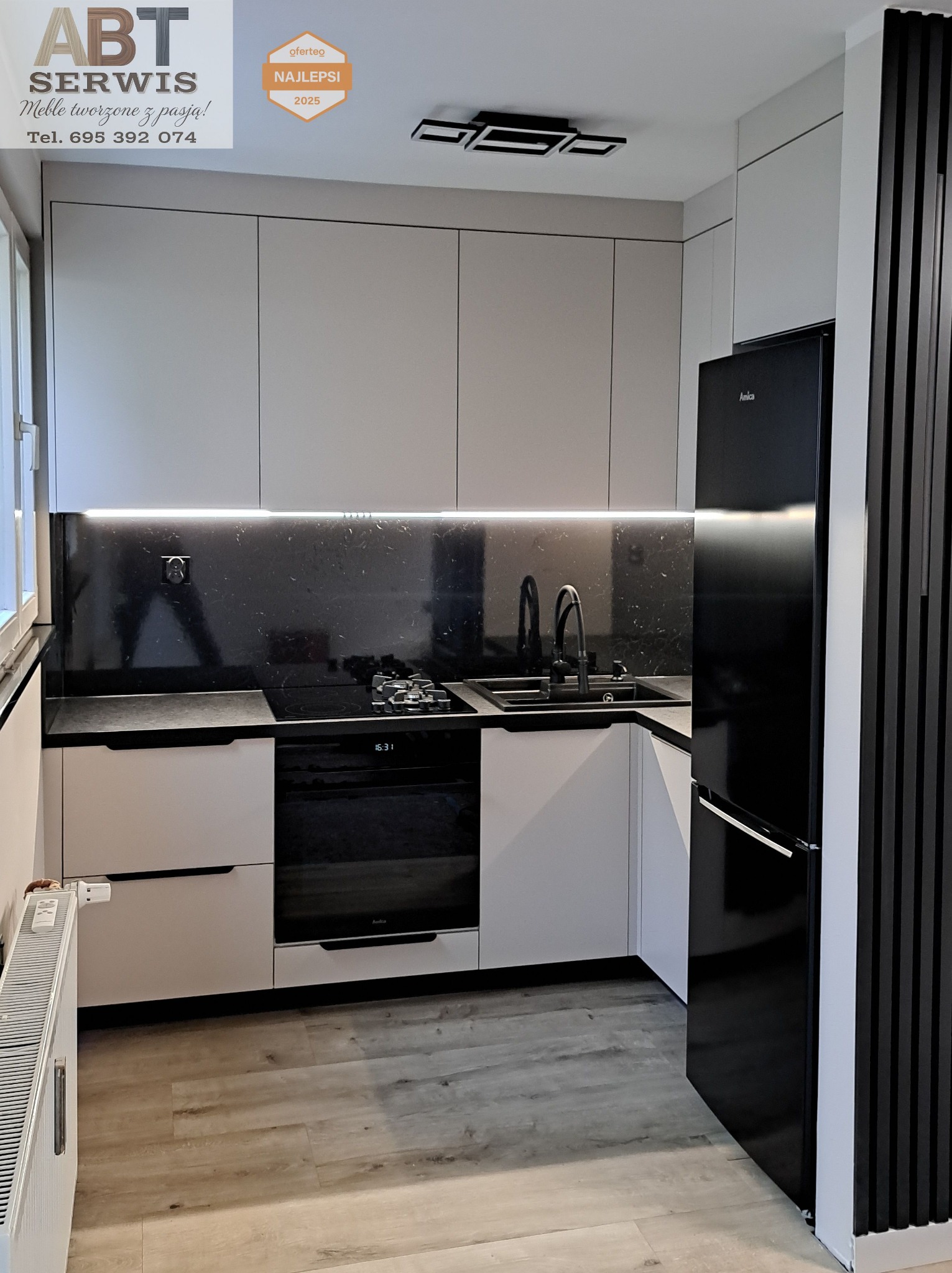 Nowoczesna kuchnia z jasnymi szafkami, czarnym blatem i lodówką Amica. Uwagę przykuwa czarny panel dekoracyjny z pionowych listew obok lodówki.