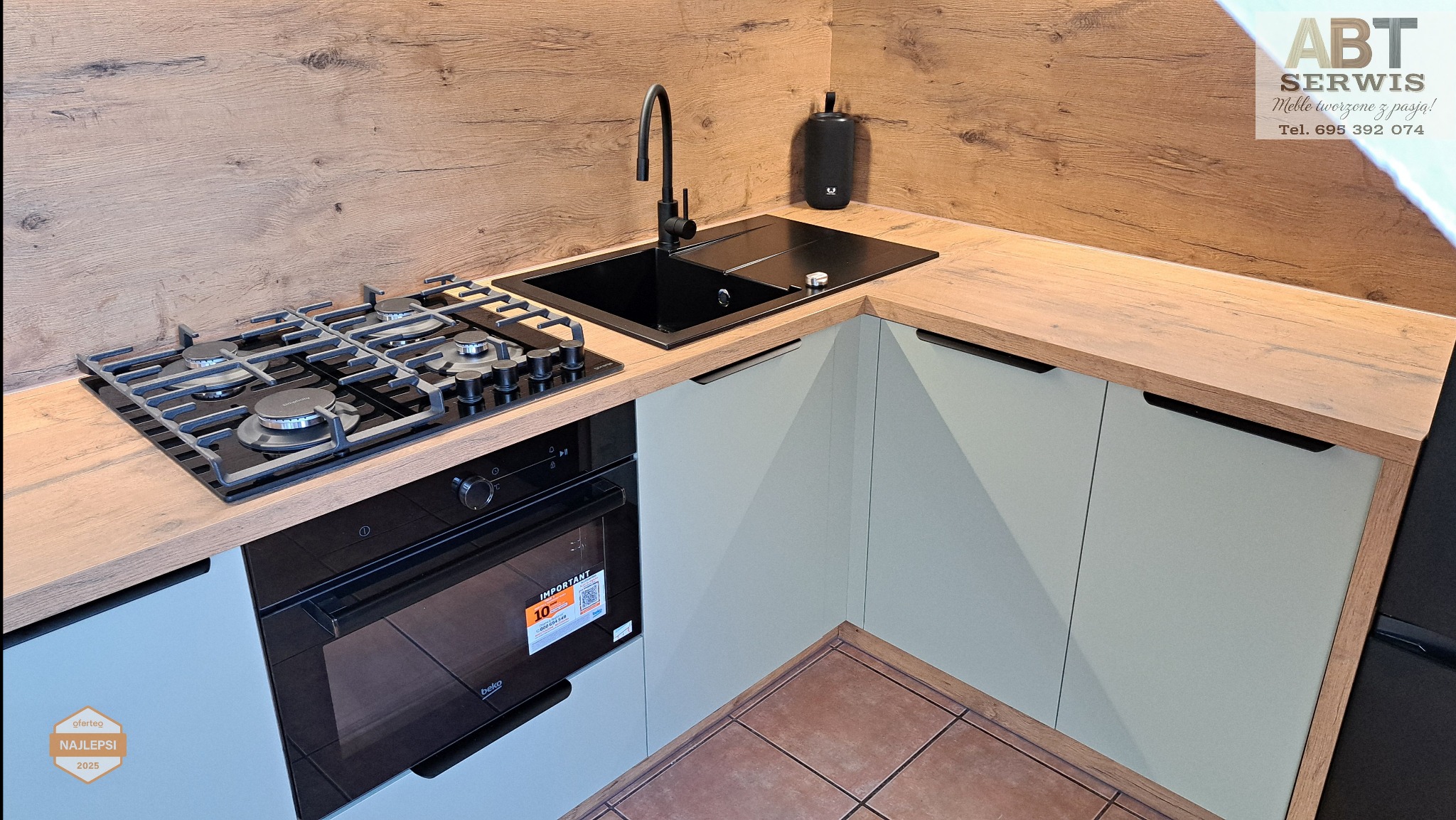 Nowoczesna kuchnia z drewnianym blatem i frontami w odcieniach szarości. Czarny zlew i płyta gazowa. Widoczny fragment podłogi z brązowych płytek.