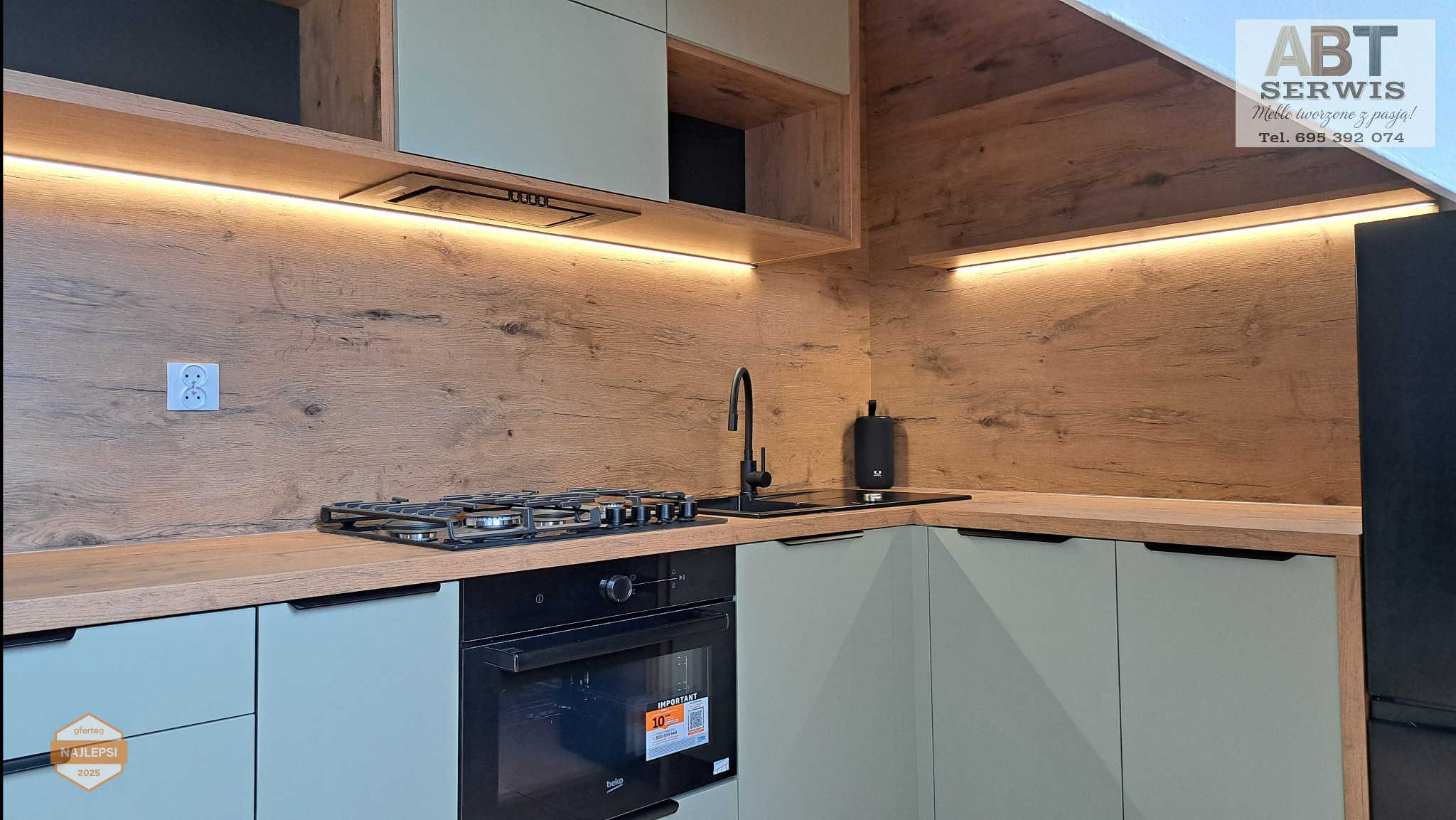 Nowoczesna kuchnia z drewnianym panelem ściennym i oświetleniem LED. Jasne szafki dolne, czarny zlew i piekarnik. Widoczny fragment lodówki i logo firmy w rogu.
