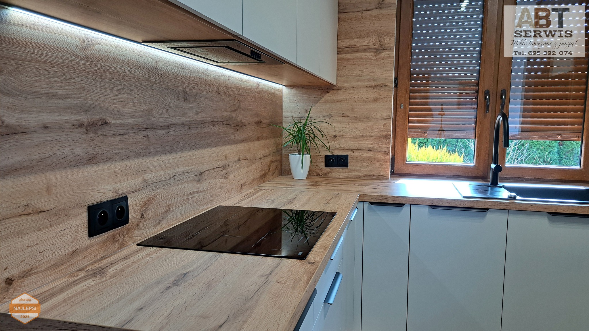 Nowoczesna kuchnia z drewnianym blatem i białymi szafkami, oświetleniem LED pod szafkami wiszącymi i czarną płytą indukcyjną. Ujęcie z bliska, widok na okno.