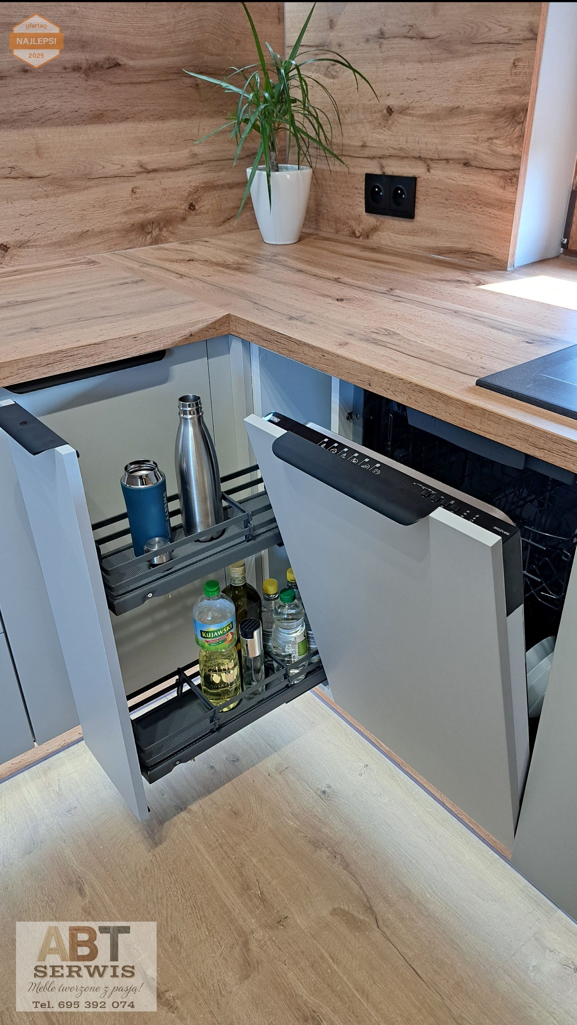 Nowoczesna kuchnia z drewnianym blatem i szarymi szafkami. Otwarta szafka cargo z butelkami oleju i napojami. Roślina w doniczce na blacie.