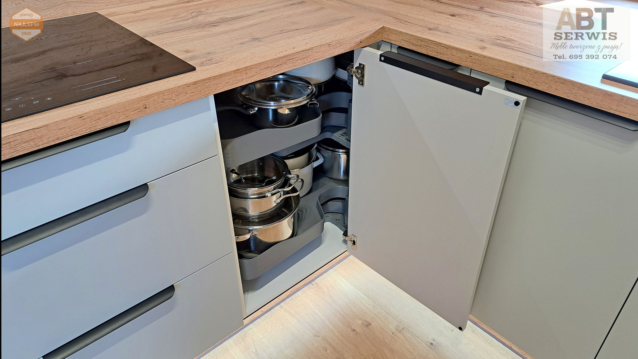 Szara kuchnia narożna z drewnianym blatem i otwartą szafką narożną z systemem półek obrotowych, na których stoją garnki. Widoczne szuflady z podłużnymi uchwytami.