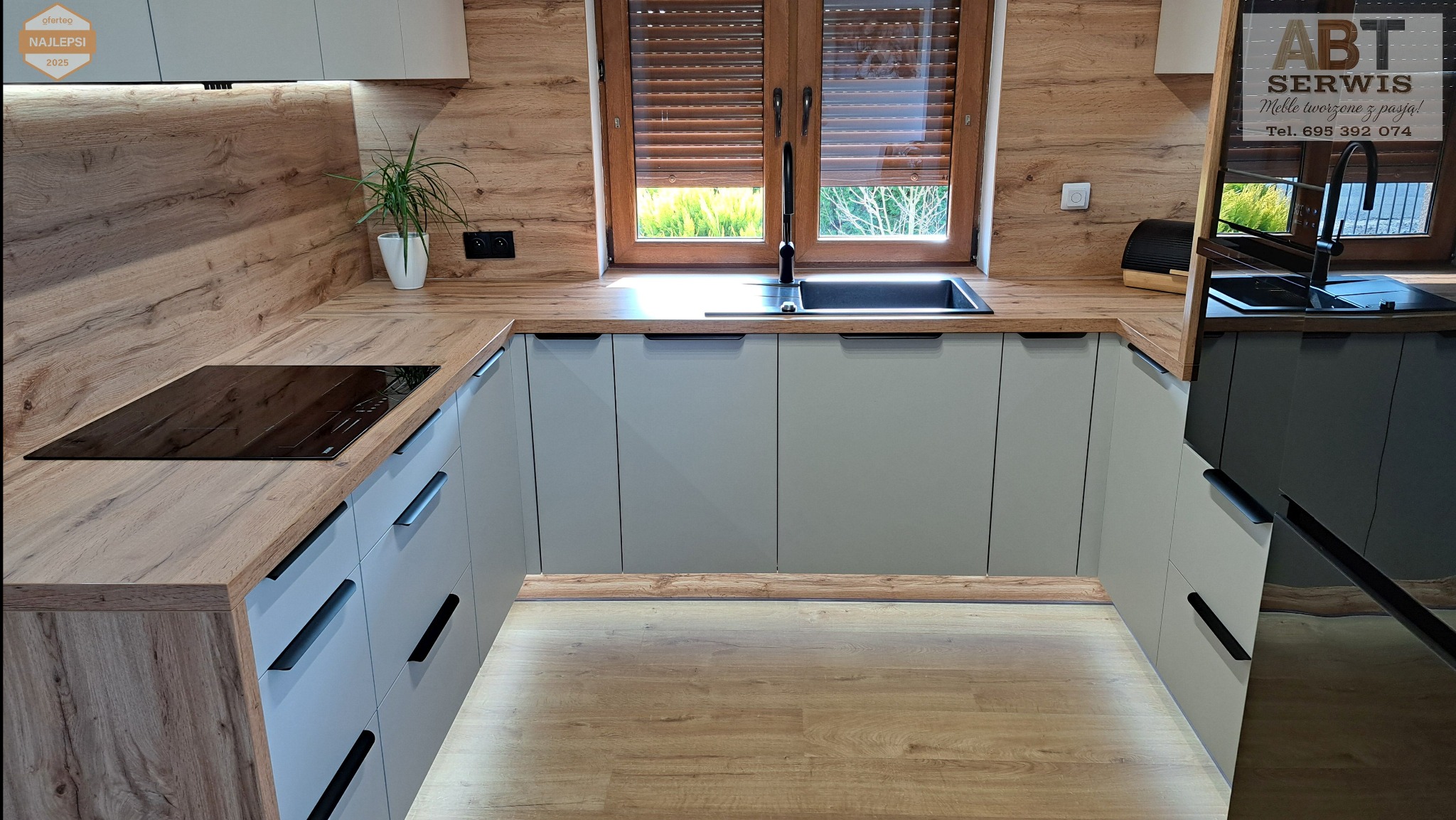 Nowoczesna kuchnia na wymiar z akrylowymi frontami w matowym wykończeniu i blatem z laminowanego dębu Wotan. Widoczna płyta indukcyjna i zlewozmywak pod oknem.