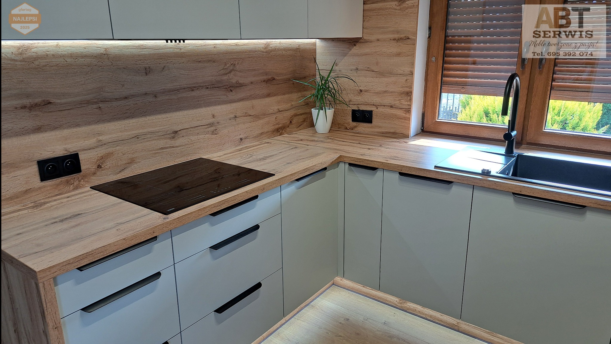 Nowoczesna kuchnia z drewnianym blatem i szarymi szafkami. Widoczna płyta indukcyjna, zlew z czarną baterią i roślina w białej doniczce. Oświetlenie LED pod szafkami.