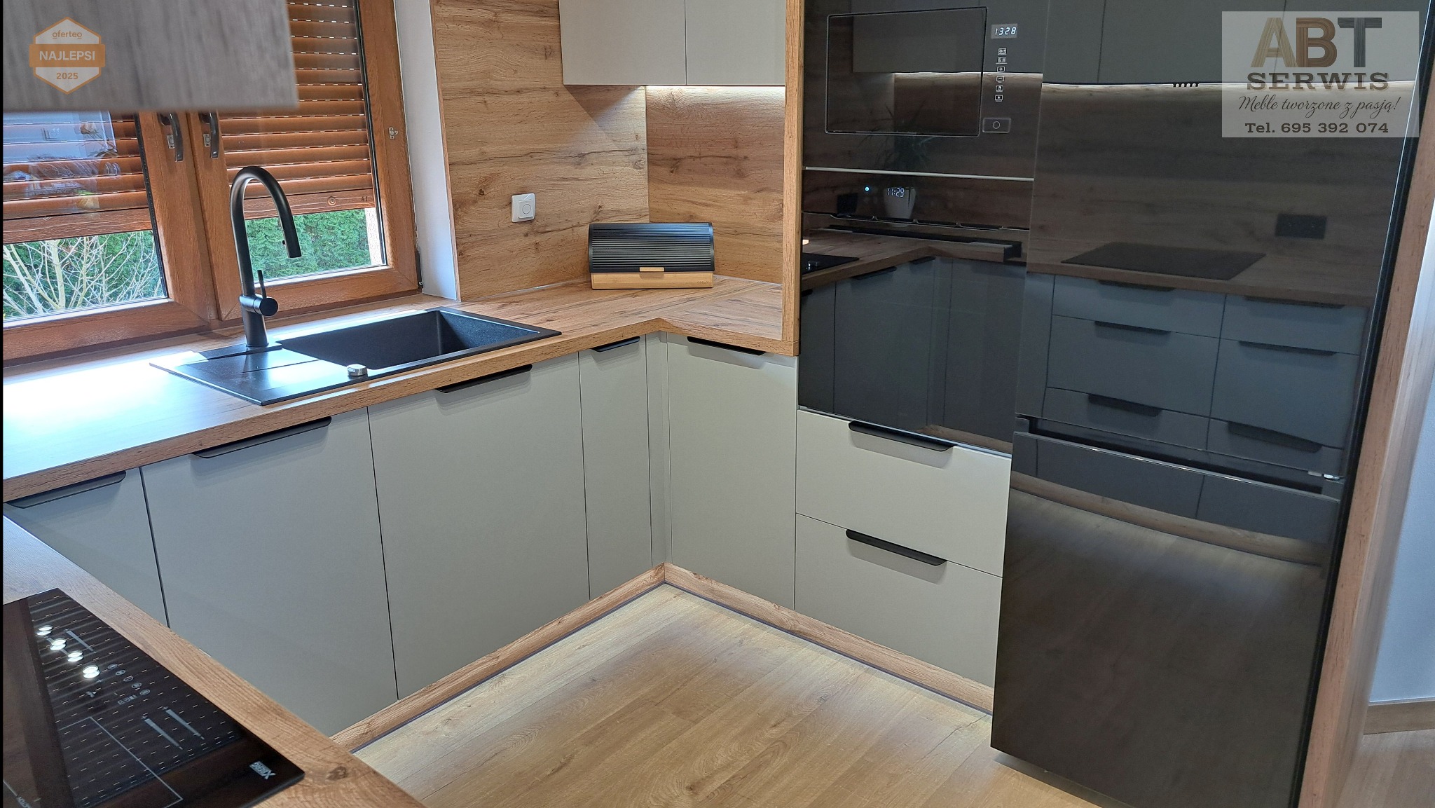 Nowoczesna kuchnia z szarymi szafkami, drewnianym blatem i okładziną. Czarny zlew pod oknem z żaluzjami. Zabudowa z piekarnikiem i lodówką w ciemnym fornirze.