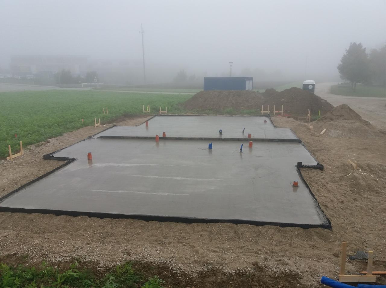 Wylany betonowy fundament płyty na gruncie, widoczne pionowe rury instalacyjne, drewniane szalunki i izolacja krawędziowa, w tle mgła i kontener budowlany.