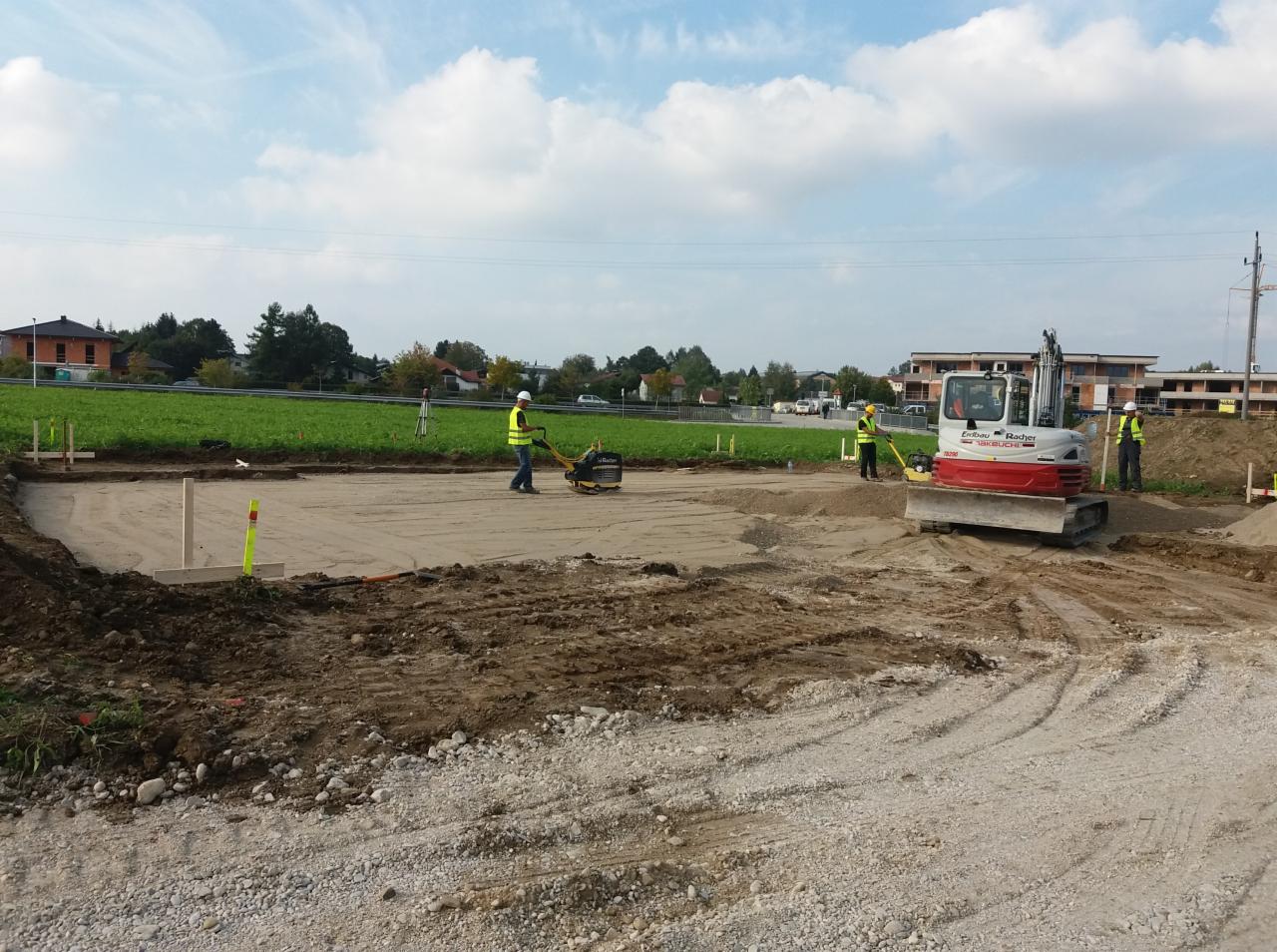 Prace ziemne pod budowę: ubijanie podłoża pod fundamenty przy użyciu zagęszczarki i mini koparki, widoczni pracownicy w kamizelkach odblaskowych, drewniane szalunki.