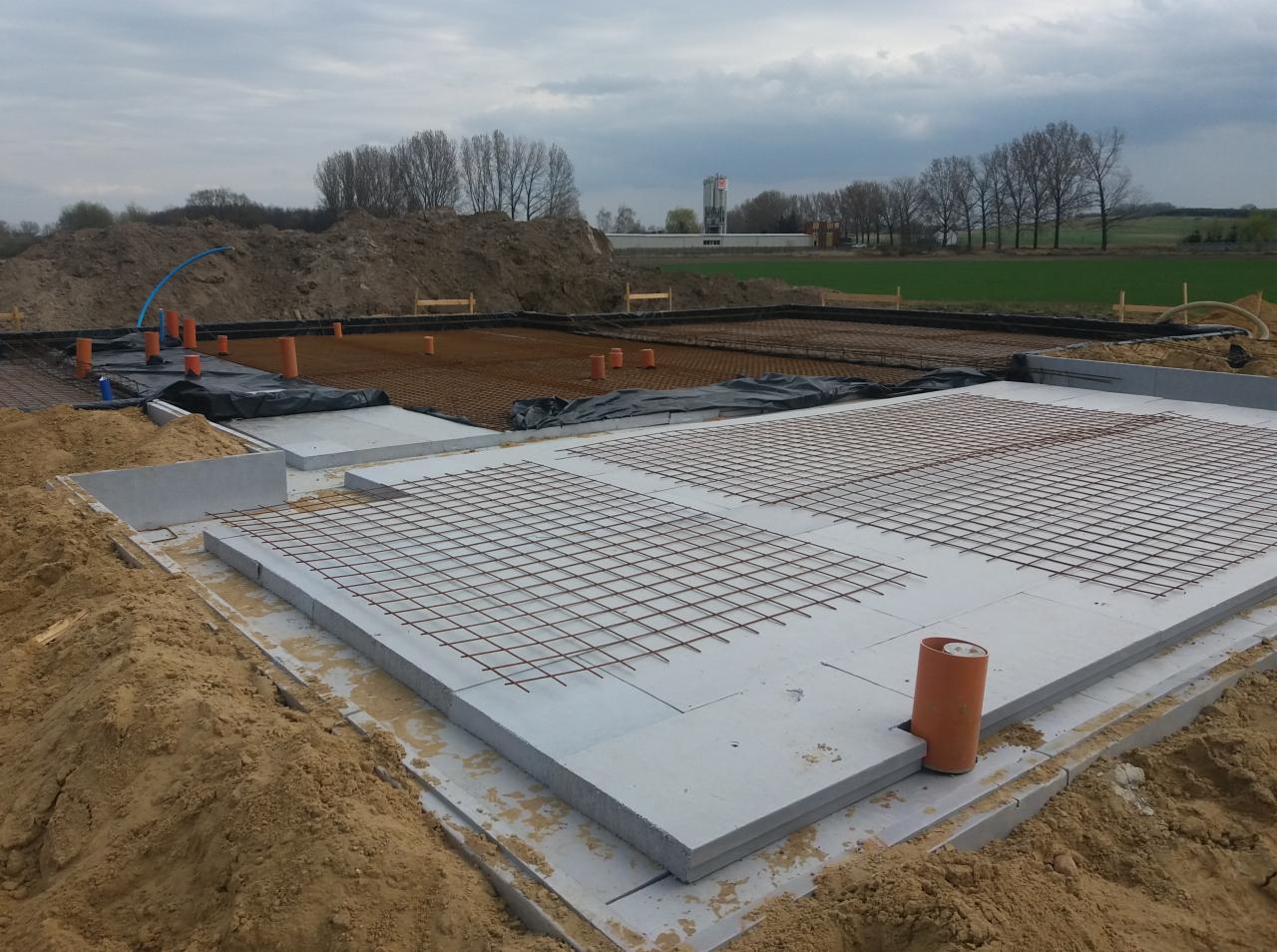 Przygotowanie pod płytę fundamentową z widoczną siatką zbrojeniową, rurami instalacyjnymi i podsypką piaskową, w tle drzewa i pochmurne niebo.