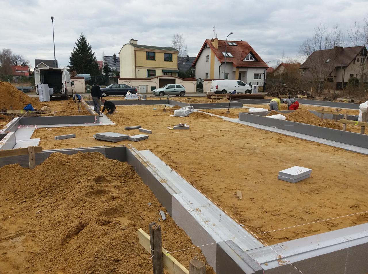 Budowa fundamentów domu jednorodzinnego: widok na wykop z szalunkami i izolacją termiczną, pracownicy w tle, hałda piasku i zabudowa jednorodzinna w tle.