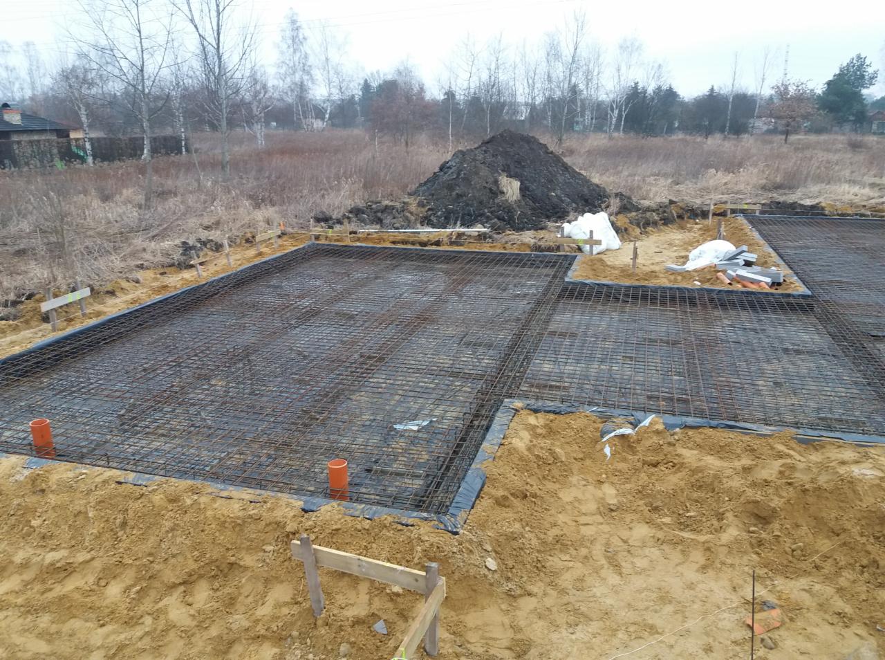 Zbrojenie fundamentów z widocznymi pomarańczowymi rurami kanalizacyjnymi, czarną folią izolacyjną i stertą ziemi w tle, wykonane w pochmurny dzień.