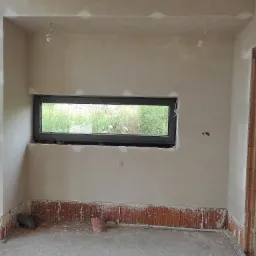 Wnętrze w trakcie prac wykończeniowych: świeżo położone tynki na ścianach, widoczne fragmenty instalacji elektrycznej, małe okno z widokiem na zieleń, ślady po pracach murarskich przy podłodze.