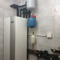 Biała pompa ciepła z widocznymi instalacjami hydraulicznymi i zbiornikami wyrównawczymi w kotłowni wykończonej płytkami w odcieniach szarości.