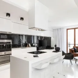 Nowoczesna kuchnia z białymi szafkami, czarnym marmurowym backsplashiem i wyspą kuchenną z białymi krzesłami barowymi, widok na salon z brązową skórzaną kanapą i czarnymi fotelami.