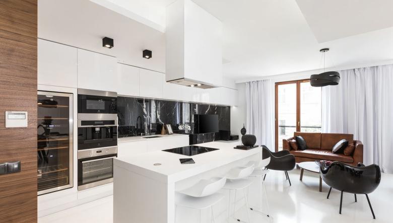 Nowoczesna kuchnia z białymi szafkami, czarnym marmurowym backsplashiem i wyspą kuchenną z białymi krzesłami barowymi, widok na salon z brązową skórzaną kanapą i czarnymi fotelami.