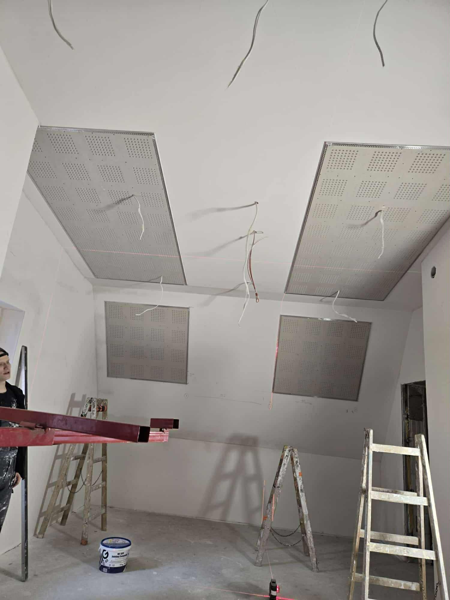 Montaż sufitu podwieszanego z płyt perforowanych w jasnym wnętrzu. Widoczne drabiny, narzędzia i wystające kable. Laserowy poziom wskazuje linię montażu.