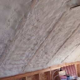 Poddasze ocieplone pianką PUR, widoczne krokwie i fragment ściany. Aplikacja pianki równomierna, pokrywająca całą powierzchnię, z detalami drewnianej konstrukcji.