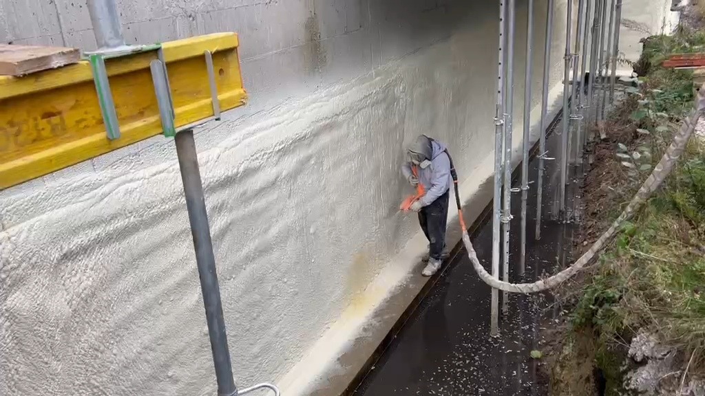 Pracownik w kombinezonie ochronnym aplikuje piankę PUR na ścianę fundamentową w Charzyno. Widoczne podpory szalunkowe i świeżo nałożona izolacja.