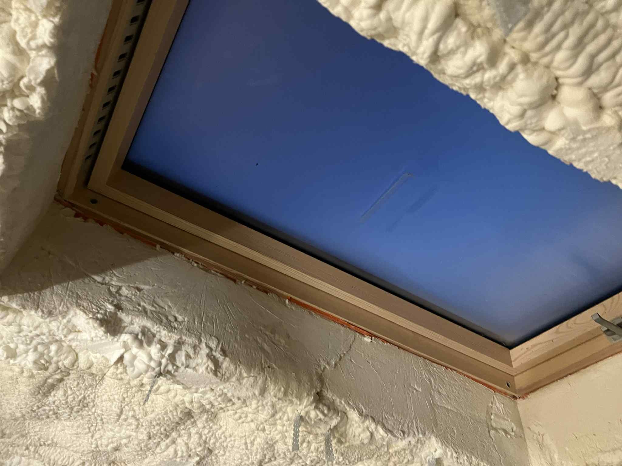 Poddasze z oknem dachowym po ociepleniu pianką PUR, widoczne detale drewnianej ramy okiennej i struktura pianki. Niebieskie niebo w tle.