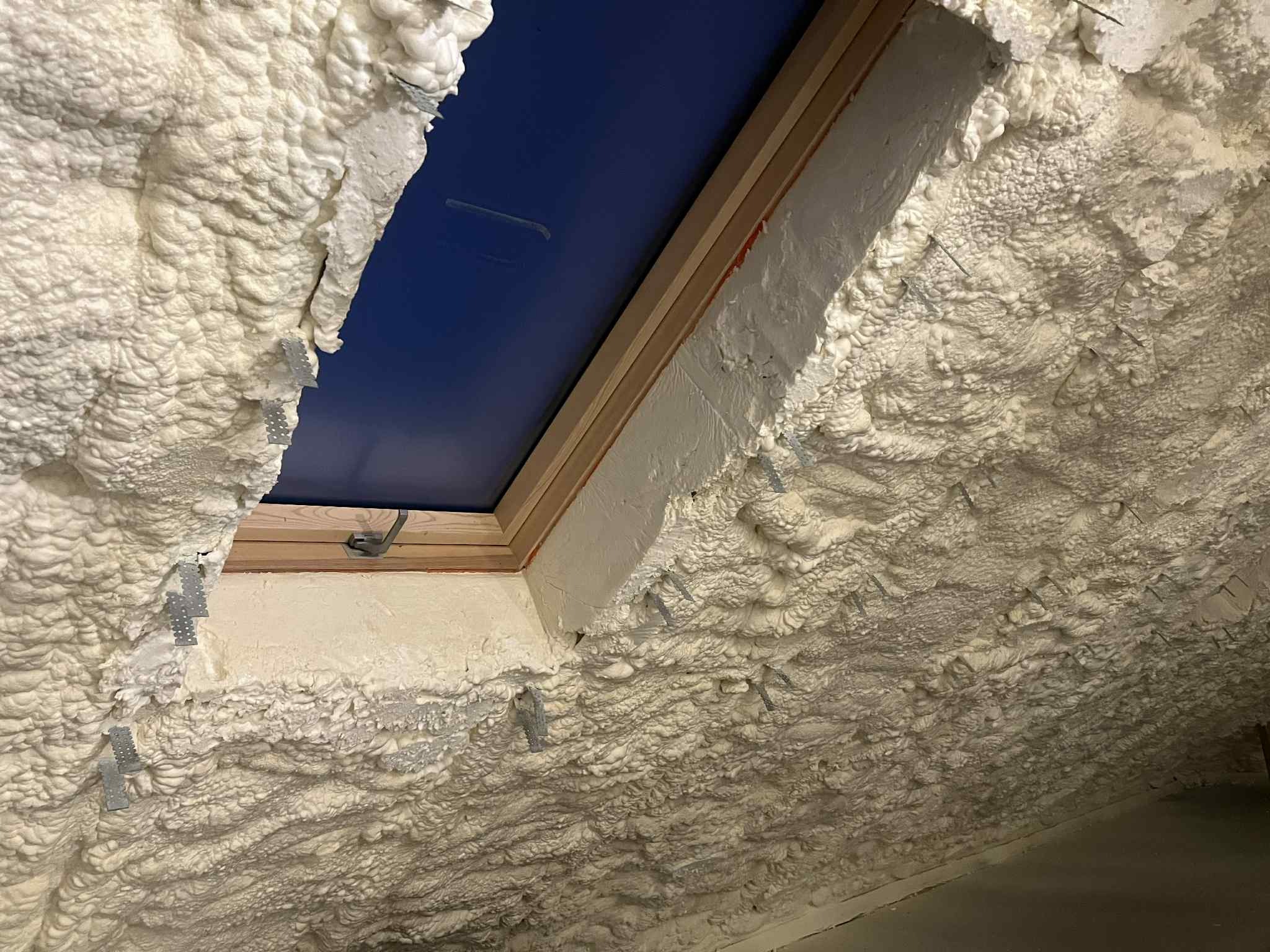 Poddasze po ociepleniu pianką PUR, widoczne metalowe mocowania i okno dachowe. Biała, nierówna struktura pianki pokrywa całą powierzchnię.