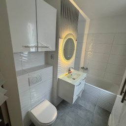 Adrian Lemke - Nowoczesna, jasna łazienka z białą armaturą, umywalką z podświetlanym lustrem i toaletą wiszącą, wykończona płytkami z falistym wzorem na ścianach i podłodze.