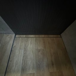 Adrian Lemke - Ukończona łazienka z podłogą wyłożoną płytkami imitującymi drewno i czarnym odpływem liniowym w rogu, widok z góry pod kątem.