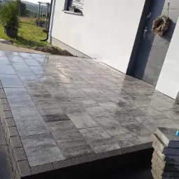 Ułożona, częściowo mokra kostka brukowa w odcieniach szarości przed wejściem do domu z białymi ścianami i szarymi drzwiami, obok drzwi wisi wieniec, w tle widoczna zieleń.