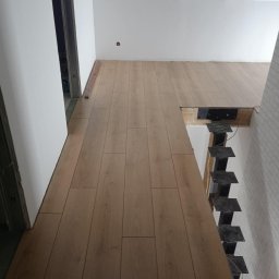 Adrian Lemke - Montaż paneli podłogowych w jasnym pomieszczeniu z widocznymi schodami szkieletowymi w tle, brak listew przypodłogowych.