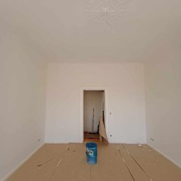 Adrian Lemke - Przygotowane do malowania pomieszczenie z zabezpieczoną podłogą i dwoma niebieskimi wiadrami z farbą na środku. W tle widoczne otwarte drzwi do kolejnego pomieszczenia.