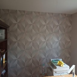 Adrian Lemke - Pokój z tapetą w geometryczne wzory 3D, widoczny regał z książkami i biurko z drukarką, butelką wody i żółtą ścierką.