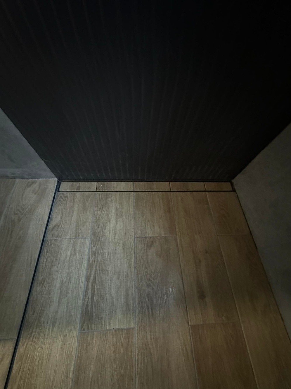 Ukończona podłoga w łazience z drewnopodobnych płytek ceramicznych i liniowym odpływem, widok z dołu, ciemny sufit z listwami.