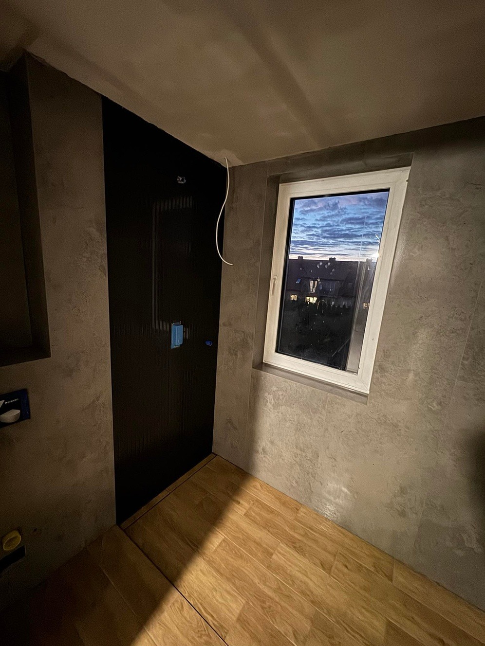 Wnętrze łazienki w trakcie remontu: szara ściana z fakturą, drewnopodobna podłoga, czarne drzwi z ukrytą ramą, okno z widokiem na zmierzch, wystający kabel elektryczny.
