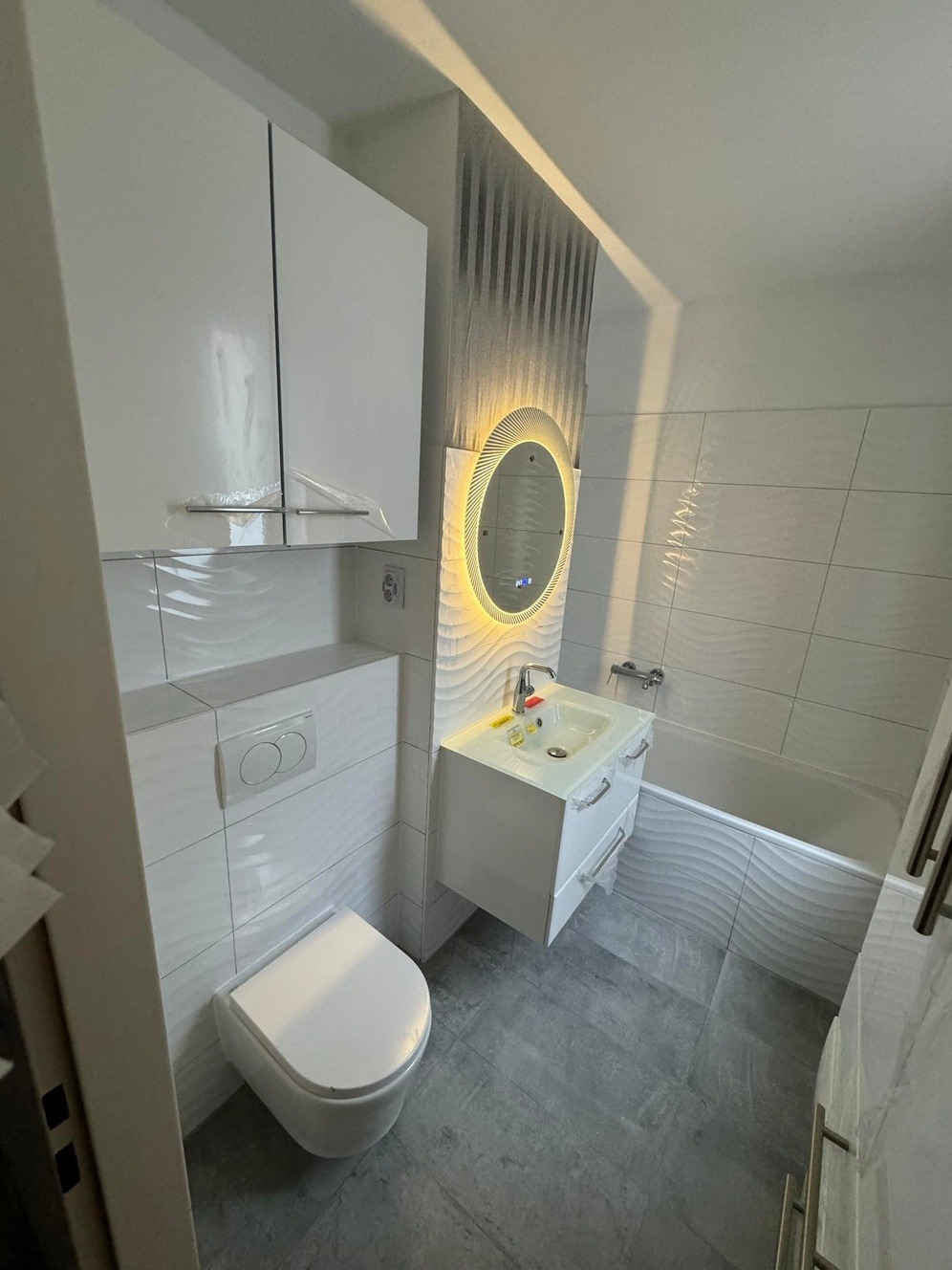 Nowoczesna, jasna łazienka z białą armaturą, umywalką z podświetlanym lustrem i toaletą wiszącą, wykończona płytkami z falistym wzorem na ścianach i podłodze.