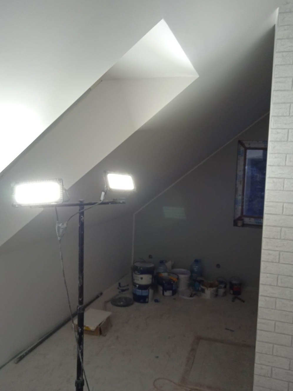 Poddasze w trakcie malowania, widoczne wiadra z farbą, lampa oświetleniowa na statywie i fragment ściany wykończonej białymi płytkami imitującymi cegłę.