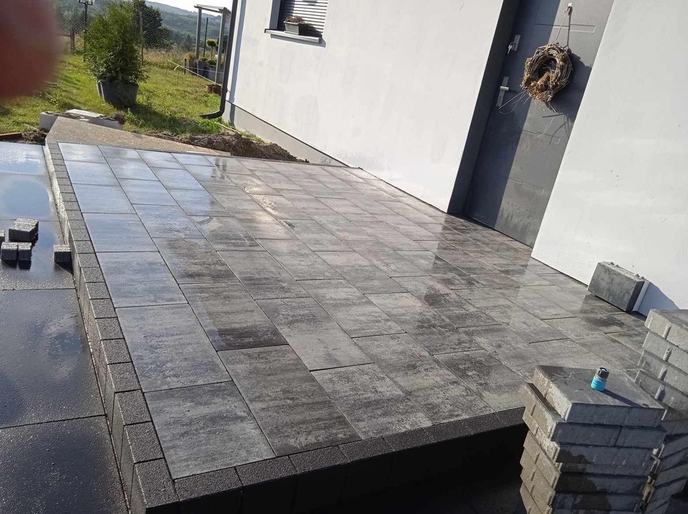 Ułożona, częściowo mokra kostka brukowa w odcieniach szarości przed wejściem do domu z białymi ścianami i szarymi drzwiami, obok drzwi wisi wieniec, w tle widoczna zieleń.