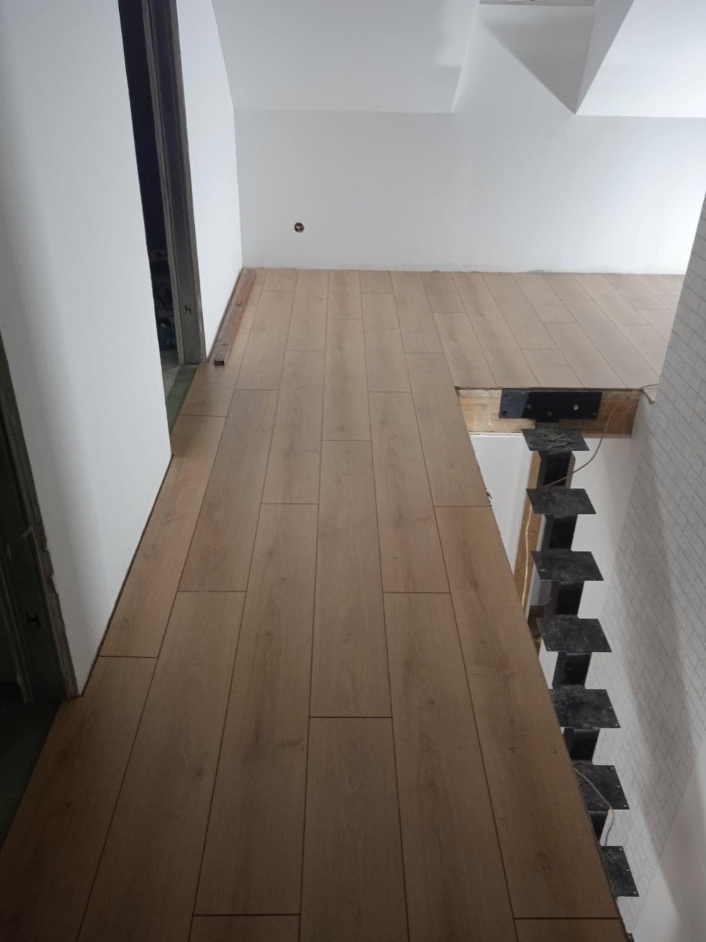 Montaż paneli podłogowych w jasnym pomieszczeniu z widocznymi schodami szkieletowymi w tle, brak listew przypodłogowych.