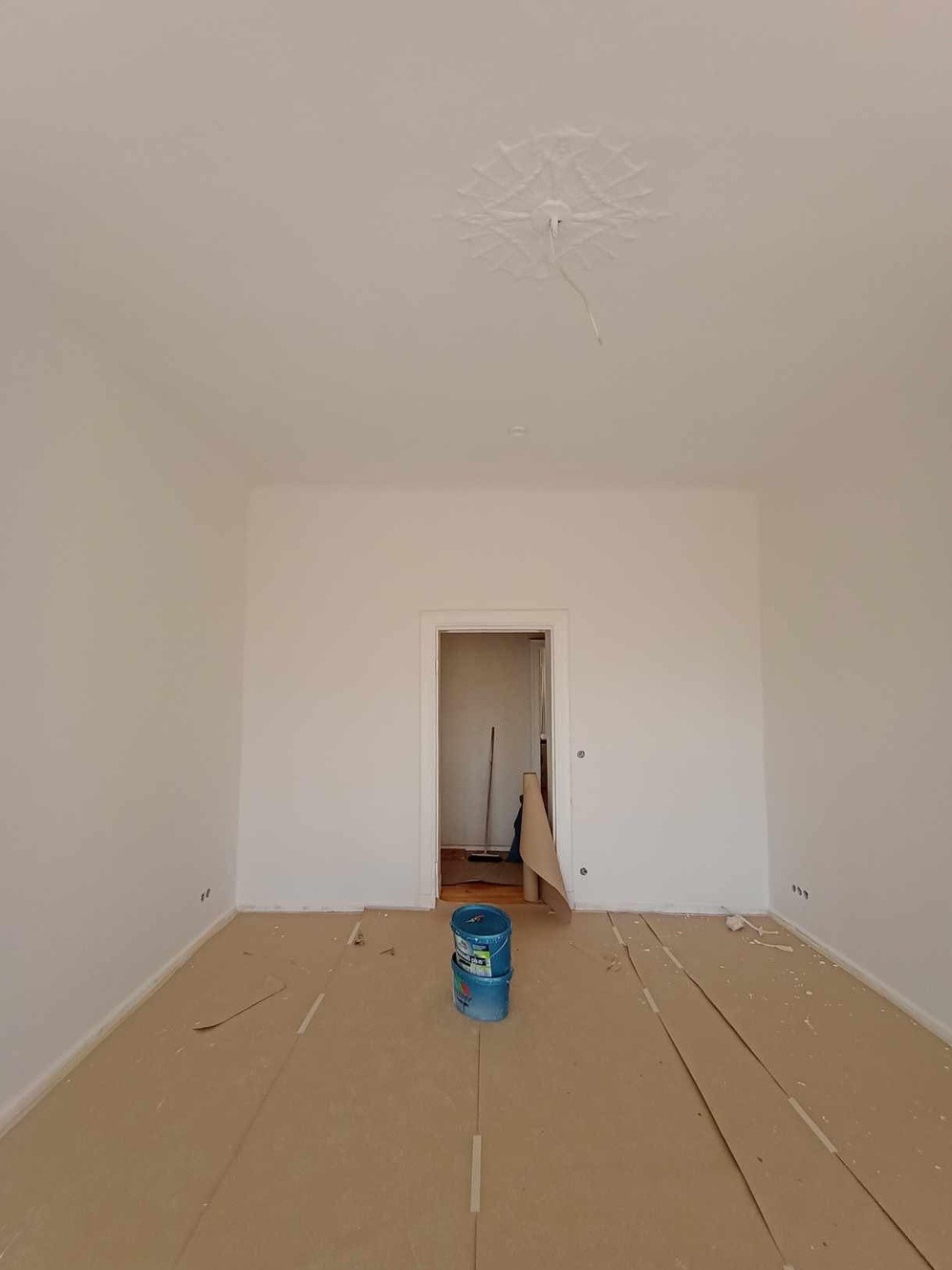 Przygotowane do malowania pomieszczenie z zabezpieczoną podłogą i dwoma niebieskimi wiadrami z farbą na środku. W tle widoczne otwarte drzwi do kolejnego pomieszczenia.