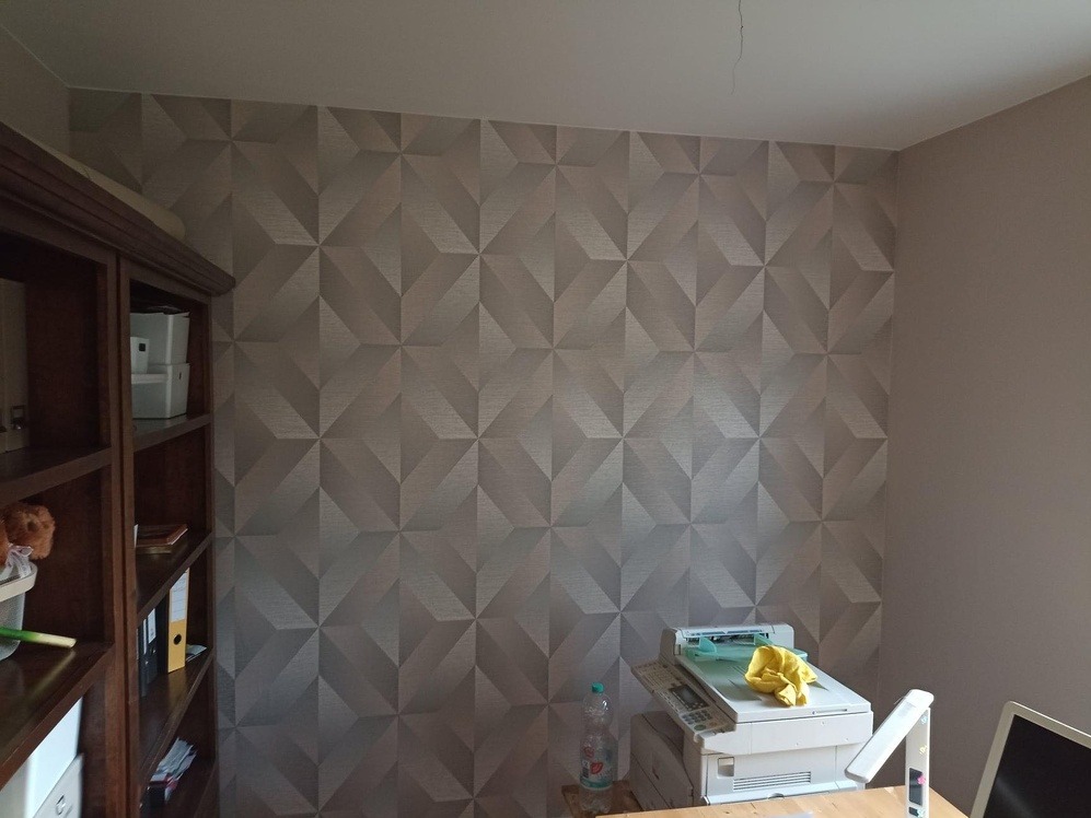 Pokój z tapetą w geometryczne wzory 3D, widoczny regał z książkami i biurko z drukarką, butelką wody i żółtą ścierką.