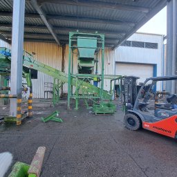 FILMAR - Montaż zielonej linii produkcyjnej w hali, widoczny wózek widłowy marki Mateco i pracownik w żółtej kamizelce, elementy konstrukcyjne na ziemi.