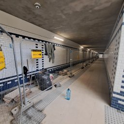 FILMAR - Wnętrze tunelu w trakcie remontu: biało-niebieska glazura metra na ścianach, tymczasowe barierki ochronne, oznakowanie 'Uwaga! Przebudowa' i 'Peron 2', butelka wody i worki z odpadami budowlanymi...