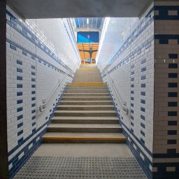 FILMAR - Schody w przejściu podziemnym wyłożone białymi i granatowymi płytkami, z żółtymi paskami antypoślizgowymi na krawędziach stopni, prowadzące ku górze w kierunku światła.