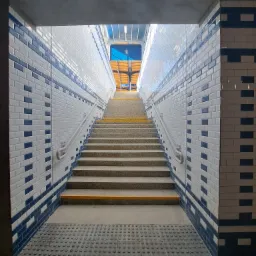 Schody w przejściu podziemnym wyłożone białymi i granatowymi płytkami, z żółtymi paskami antypoślizgowymi na krawędziach stopni, prowadzące ku górze w kierunku światła.