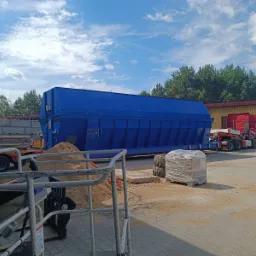 Niebieski kontener przemysłowy na przyczepie, widok z bliska z paletą zafoliowaną obok, na placu z kostki brukowej, w tle budynek i las.