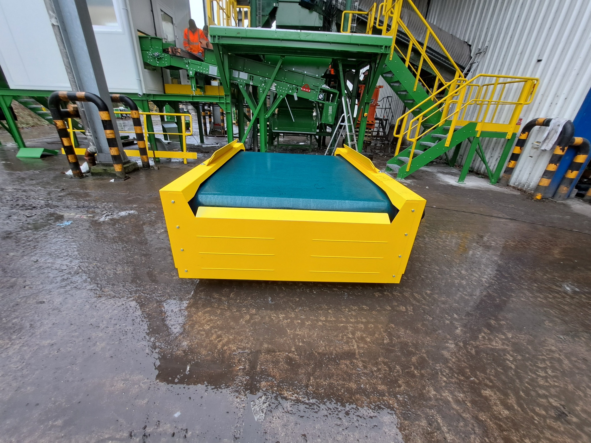 Żółty podajnik taśmowy z niebieską taśmą na mokrym placu, w tle konstrukcja stalowa z zielonymi schodami i elementami.