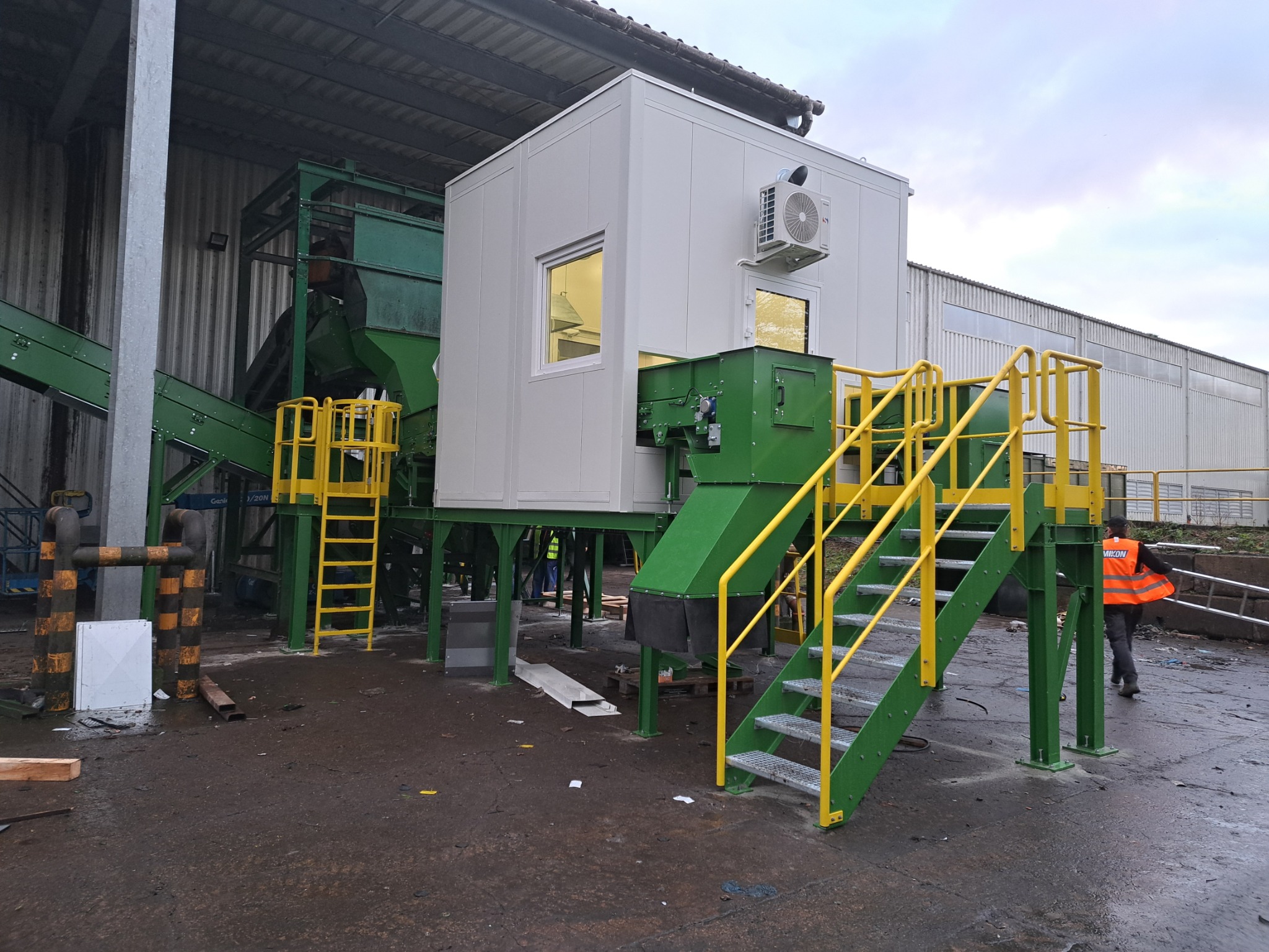Instalacja przemysłowa z zielonymi elementami konstrukcyjnymi, żółtymi schodami i białą kabiną z klimatyzatorem na zewnątrz, na tle hali przemysłowej.