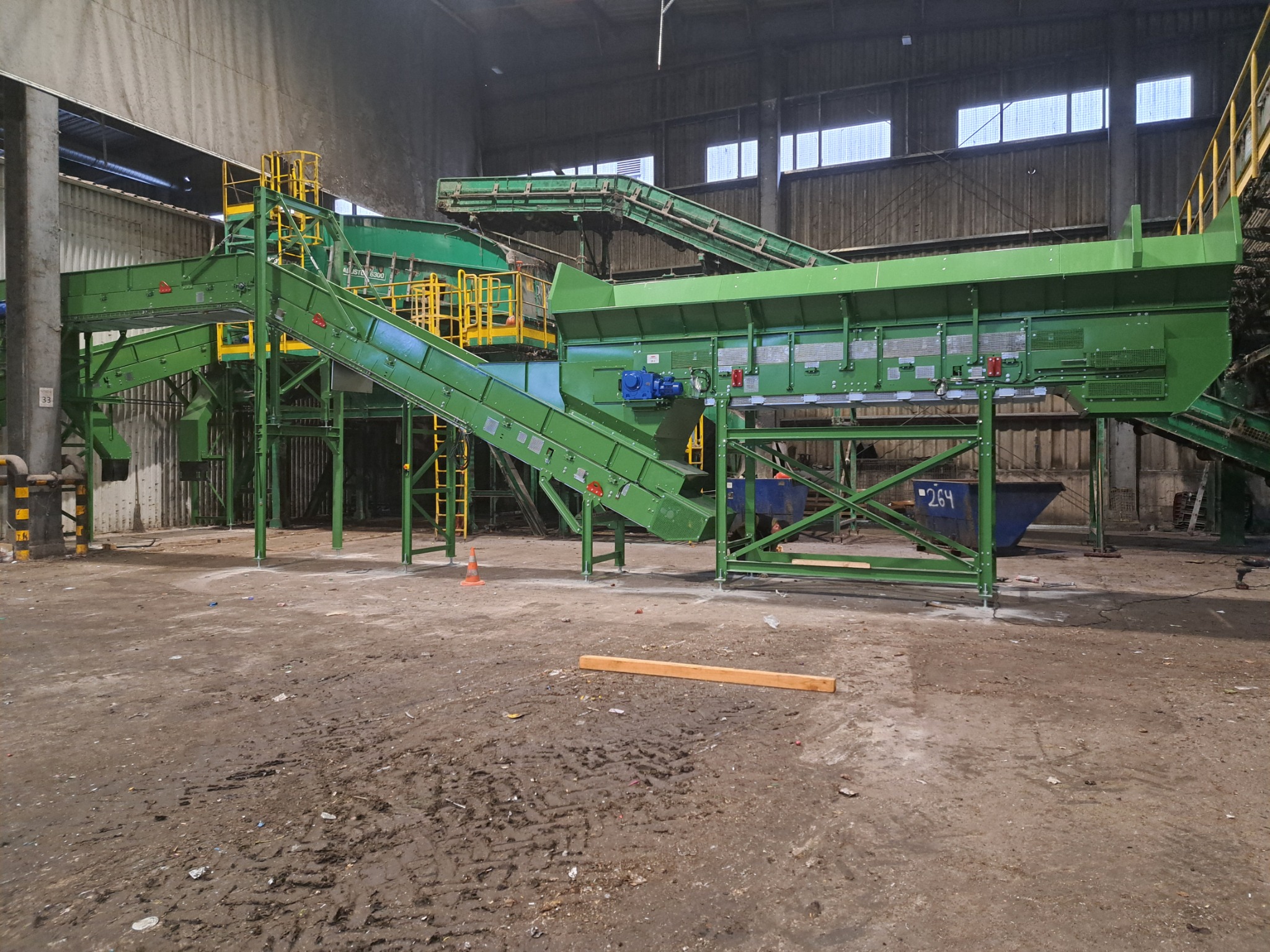 Kompleksowy system przenośników taśmowych w hali przemysłowej, pomalowany na zielono z żółtymi elementami, z niebieskim silnikiem elektrycznym, ustawiony na zabrudzonej posadzce.