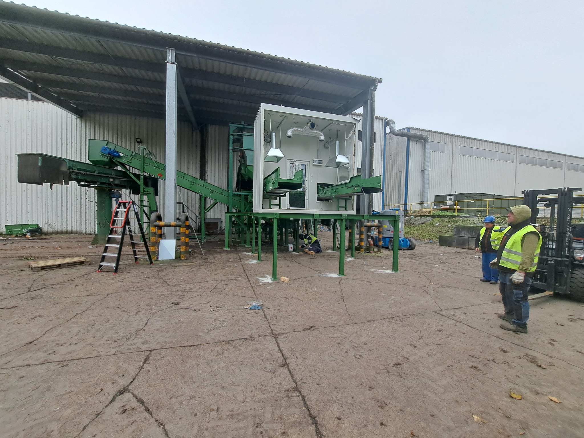 Montaż linii sortowniczej w hali przemysłowej, widoczni pracownicy w kamizelkach odblaskowych, w tle wózek widłowy.