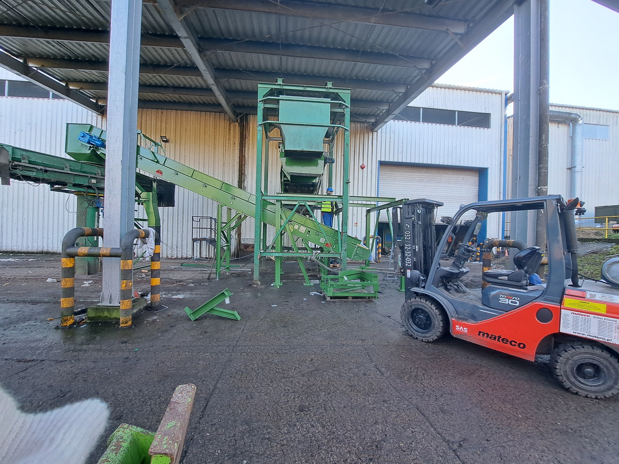 Montaż zielonej linii produkcyjnej w hali, widoczny wózek widłowy marki Mateco i pracownik w żółtej kamizelce, elementy konstrukcyjne na ziemi.