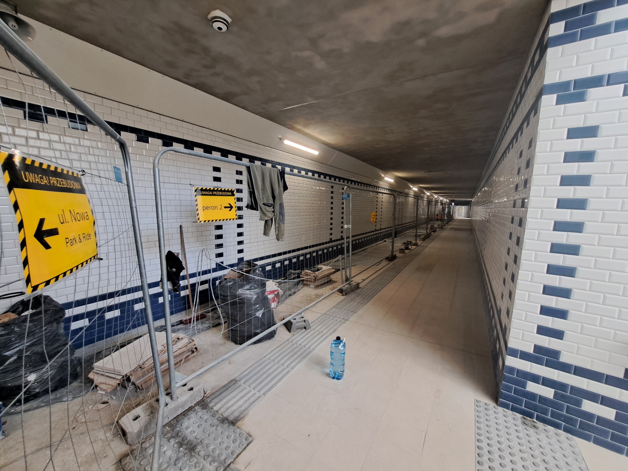 Wnętrze tunelu w trakcie remontu: biało-niebieska glazura metra na ścianach, tymczasowe barierki ochronne, oznakowanie 'Uwaga! Przebudowa' i 'Peron 2', butelka wody i worki z odpadami budowlanymi...
