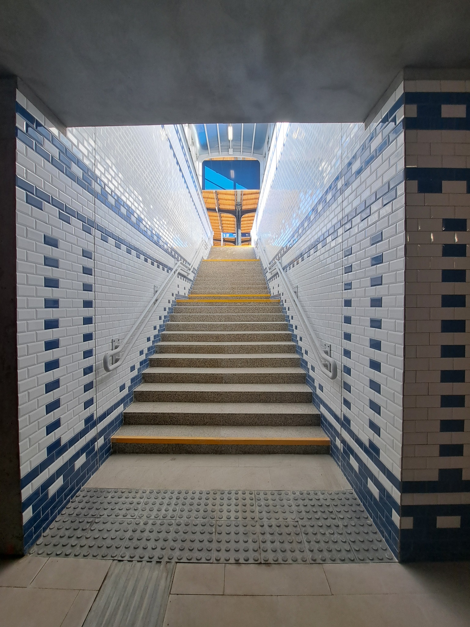 Schody w przejściu podziemnym wyłożone białymi i granatowymi płytkami, z żółtymi paskami antypoślizgowymi na krawędziach stopni, prowadzące ku górze w kierunku światła.